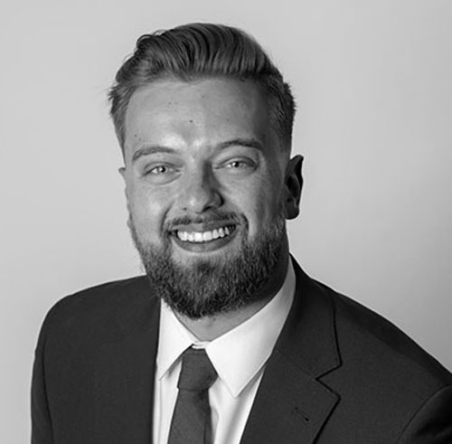 A man with a beard and short hair, wearing a suit and tie, smiles at the camera against a plain background in a black and white portrait.