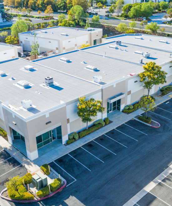 Aerial view of a modern, beige commercial building with large windows, a flat roof, and surrounding parking lot. Several empty parking spaces and landscaped areas with trees and bushes are visible.