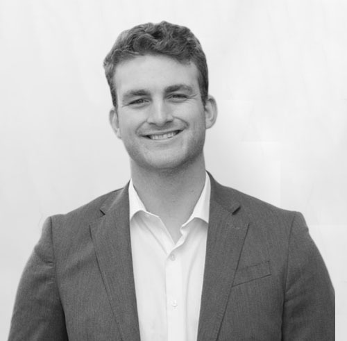 A man in a suit jacket and white shirt smiles at the camera. The background is plain and light-colored. The image is in black and white.