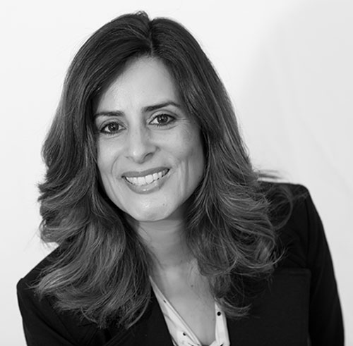 A woman with medium-length wavy hair smiles at the camera. She is wearing a dark blazer over a light-colored top, and the photo is in black and white with a plain background.
