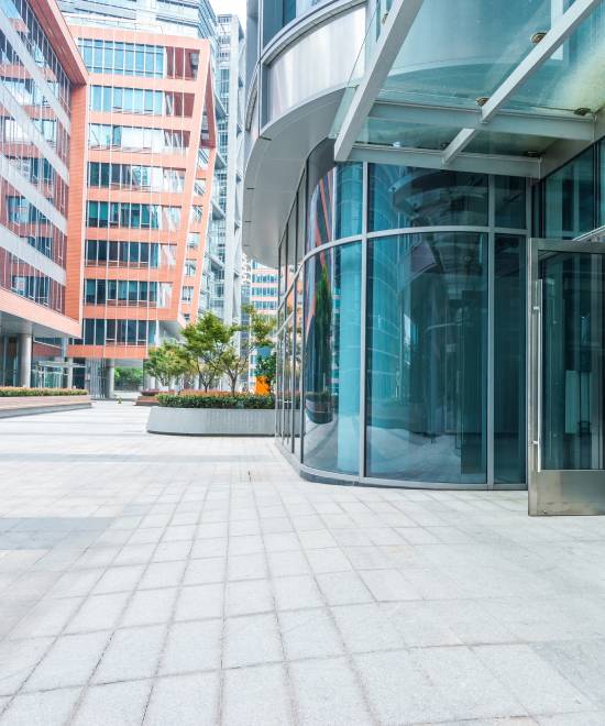 Modern glass office buildings with reflective windows line a clean, empty plaza with tiled pavement and some greenery, creating an urban business district atmosphere on a clear day.