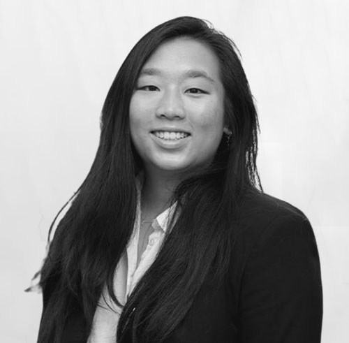 A woman with long dark hair, wearing a blazer over a light-colored shirt, smiles at the camera against a plain light background.