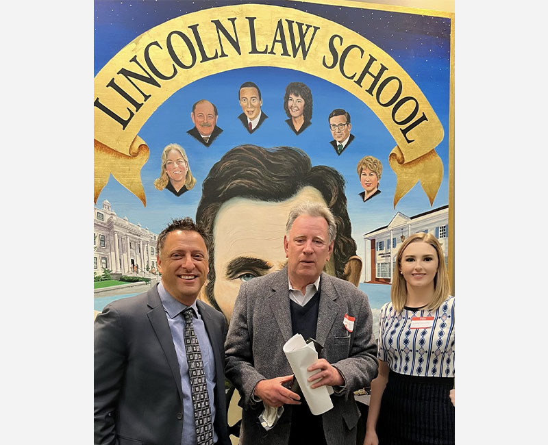 Three people stand in front of a mural featuring Abraham Lincoln and portraits of other individuals, with the text "Lincoln Law School" above them. The man in the center holds rolled papers.