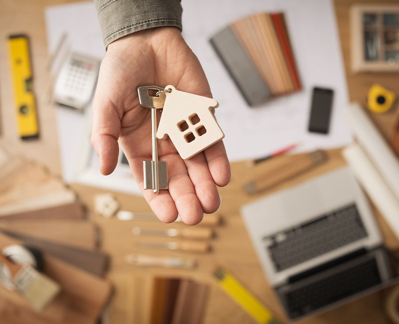A person’s hand holding a key with a house-shaped keychain, above a desk with architectural plans, wood samples, a calculator, and various construction tools.