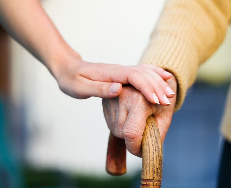 A younger hand gently resting on top of an older person's hand, which is holding a wooden cane, symbolizing support and care.
