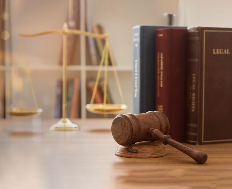 A wooden judge's gavel rests on a desk next to thick legal books, with a blurred golden balance scale and bookshelf in the background.