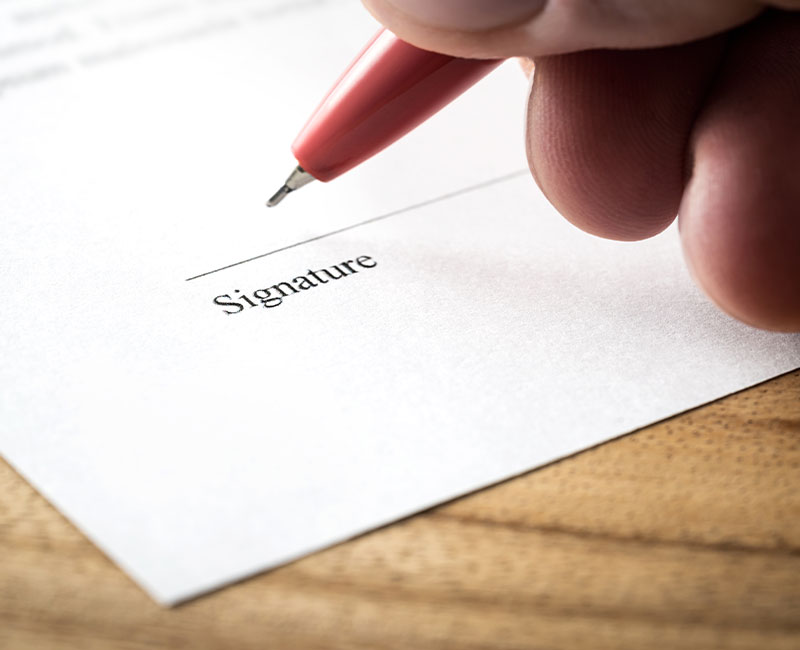 Close-up of a hand holding a pen, about to sign a document on a signature line labeled "Signature." The paper is white and the pen has a red body.