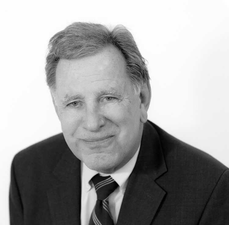 A grayscale portrait of an older man with short hair, wearing a suit, dress shirt, and striped tie, smiling gently against a plain light background.