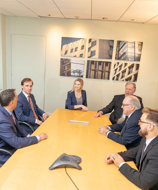 Six people in business attire sit around a conference table in a meeting room, engaged in conversation. Modern artwork featuring buildings hangs on the wall behind them. A conference phone is on the table.