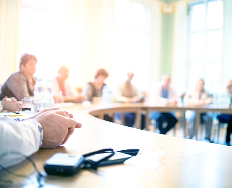 A group of people sits around a curved conference table in a bright room, with hands and electronic devices visible in the foreground. The background is out of focus, emphasizing a meeting or discussion setting.