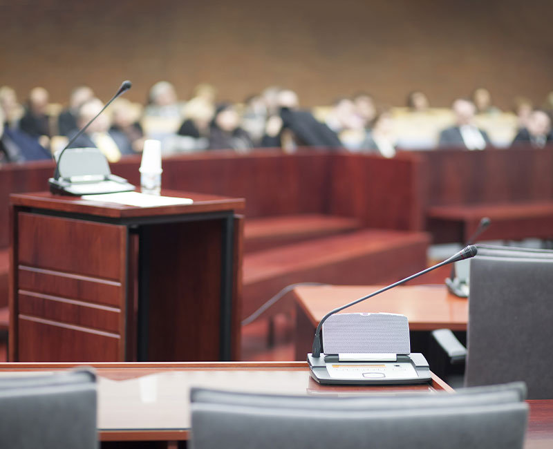 A close-up of a courtroom with microphones on desks, a wooden podium in the foreground, and blurred people seated in the background, suggesting a formal legal proceeding or trial in session.