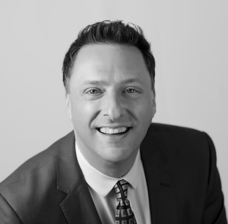 Black and white portrait of a man in a suit and patterned tie, smiling at the camera against a plain background.