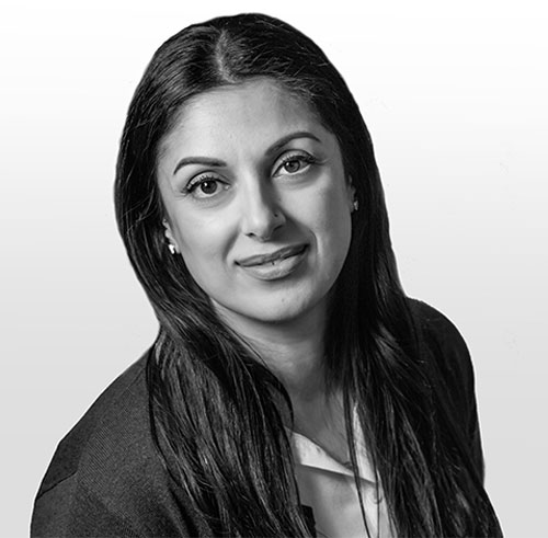 A woman with long dark hair, wearing a blazer and light-colored blouse, looks at the camera and smiles slightly. The photo is in black and white with a plain, light background.