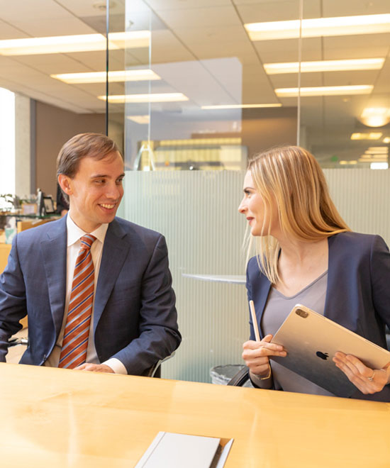Two people in business attire sit at a conference table in a bright office, smiling and talking. The woman holds a tablet and a pen, while the man looks at her, both appearing engaged in conversation.