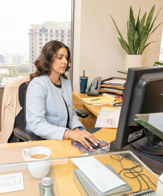 A woman in a light blue blazer works at a desk with a computer in a modern office. Papers, a plant, a coffee cup, and office supplies are on the desk, with city buildings visible through the window behind her.