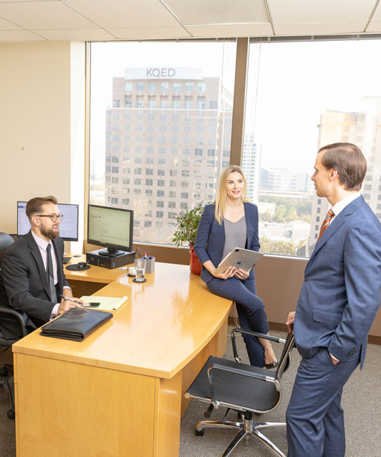 Three professionals in business attire have a discussion in a modern office with large windows. One man sits at a desk with a computer, a woman sits on the desk holding a tablet, and another man stands facing them.