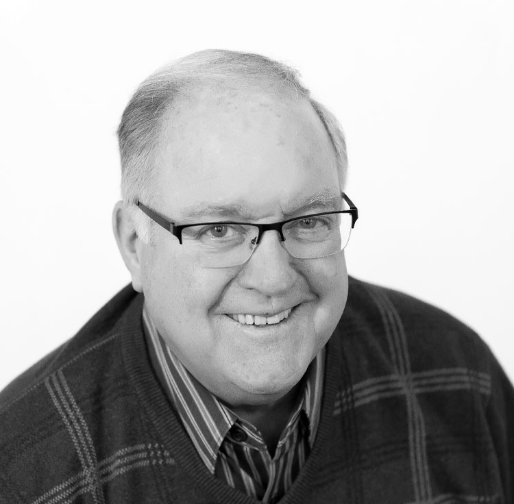 A smiling older man with short gray hair and glasses, wearing a striped shirt and a dark plaid sweater, poses against a plain, light background.