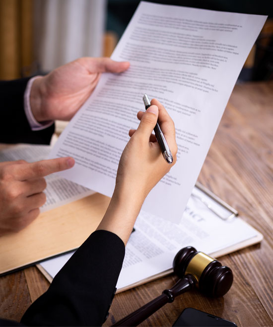 Two people review a legal document at a wooden desk, one pointing at the paper while the other holds a pen. A gavel and an open book are visible on the desk.
