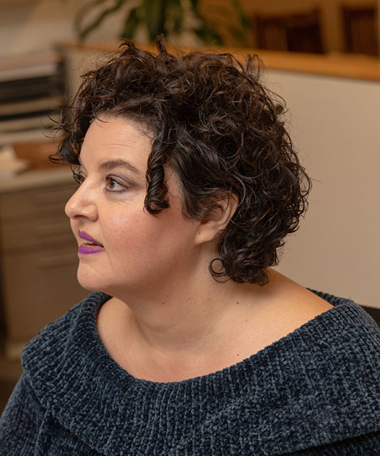 Woman with short, dark, curly hair and purple lipstick, wearing a textured dark blue off-the-shoulder top, sitting indoors and looking to the left.