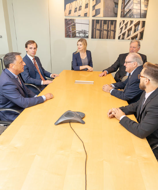 Six people in business attire sit around a long conference table in a meeting room, engaged in discussion. Modern building photos hang on the wall behind them. A conference phone is on the table.