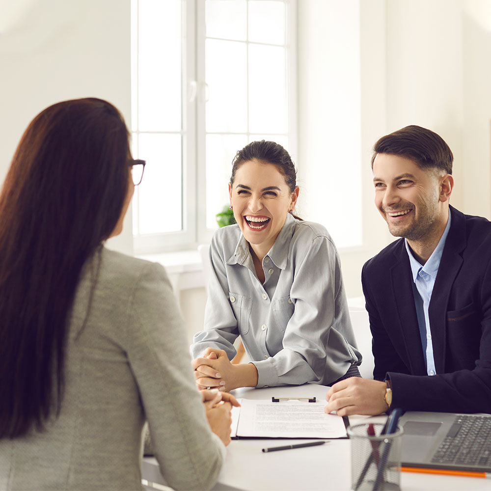 Three people sit at a desk in a bright office, facing each other. Two of them, a woman and a man, are smiling and laughing, while a woman with long dark hair sits across from them. Papers and a laptop are on the desk.