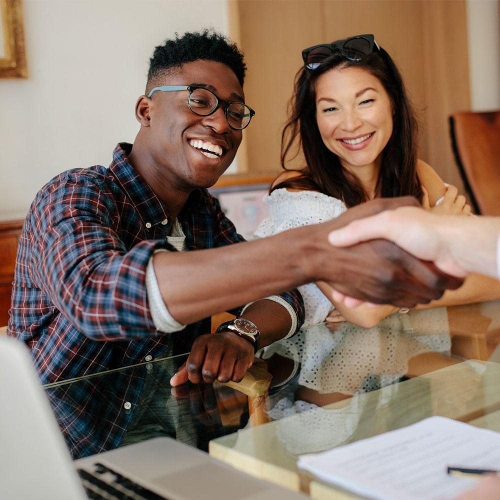 A smiling man wearing glasses shakes hands with someone across a desk, while a smiling woman sits beside him. Papers and a laptop are on the table, suggesting a business meeting or agreement.