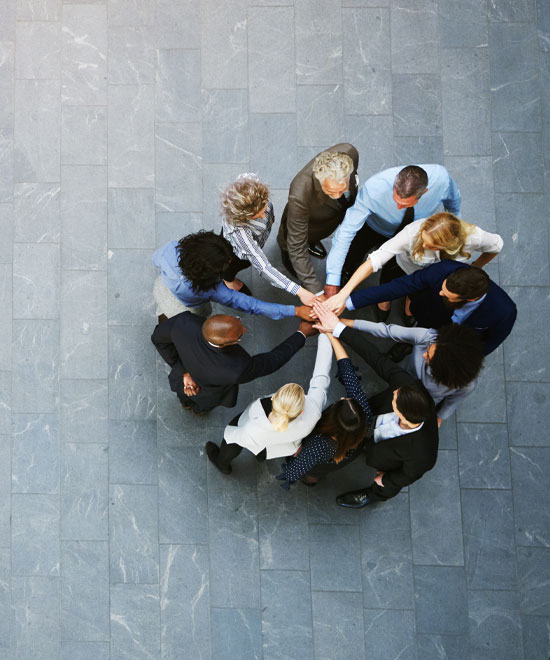 A group of people standing in a circle on a gray tiled floor, placing their hands together in the center, symbolizing teamwork and unity. Photo is taken from above.
